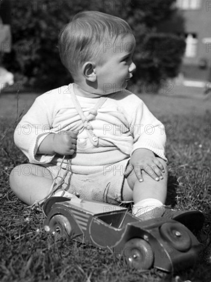 Little boy playing with a wooden car, 1930s, Toddler sitting in the grass and playing with a red toy car. It looks to the side, historical photo