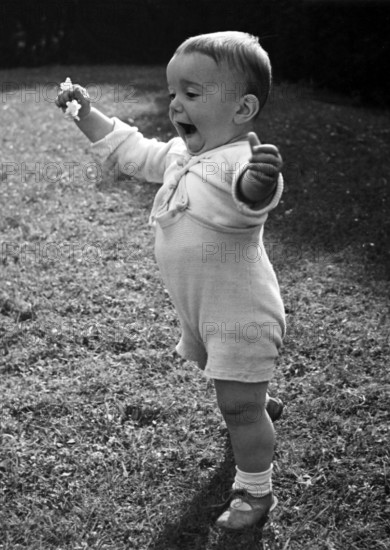 Little boy learning to walk, 1930s, Toddler stands happily in the meadow and raises his right arm, Historical photo