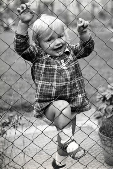 Little girl climbing over the fence, 1930s, child in a chequered dress, holding on to the fence, looking curiously through the net, historical photo