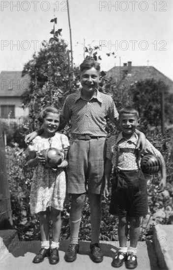 Siblings with big brother, 1950s, Three children posing with balls in a garden and smiling at the camera, historical photo