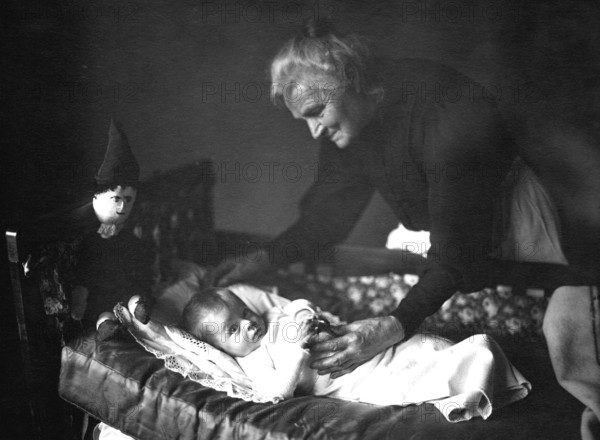 Grandma with grandson, 1930s, An elderly woman lovingly cares for a baby in a cradle, Historical photo