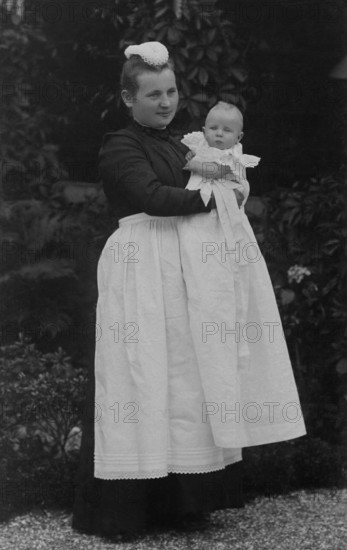 Nanny holding a baby in her arms, ca. 1910, Woman in Victorian dress holding a baby in the garden, both elegantly dressed, historical photo