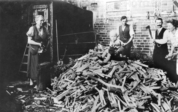 Baby sitting on a mountain of firewood, 1930s, family working together in the yard chopping wood while a child sits on the wood, historical photo