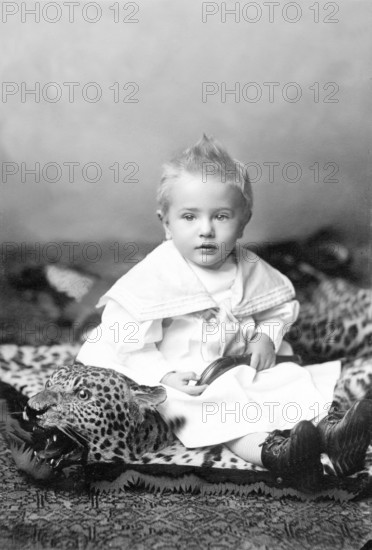 Little boy sitting on a leopard skin, 1910s, A little boy in a traditional outfit sits on a toy lion and looks into the camera, Historical photo