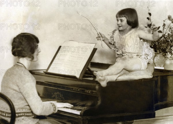 Mother with child at the piano, 1920s, A girl sits happily on a piano while a woman plays, Historical photo