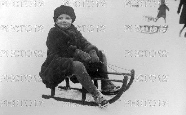 Child sledging, 1930s, A child sits on a sledge in the snow with a joyful expression on his face. Winter clothing and surroundings, Historical photo