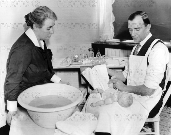 Man practising handling a baby with a doll, 1940s, A man and a woman caringly looking after a baby, next to them a basin of water, serious mood, Historical photo