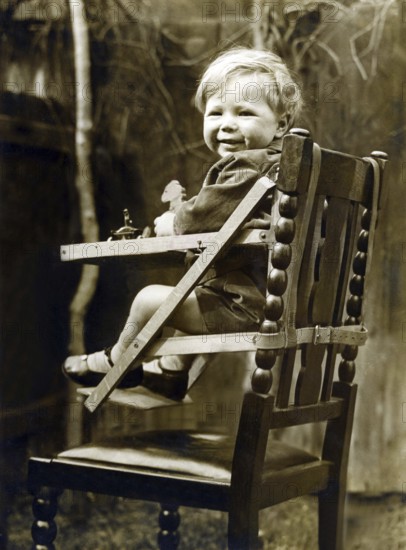 Baby in a child seat, 1930s, A smiling toddler sits happily in a wooden high chair, outside in a garden area, Historical photo