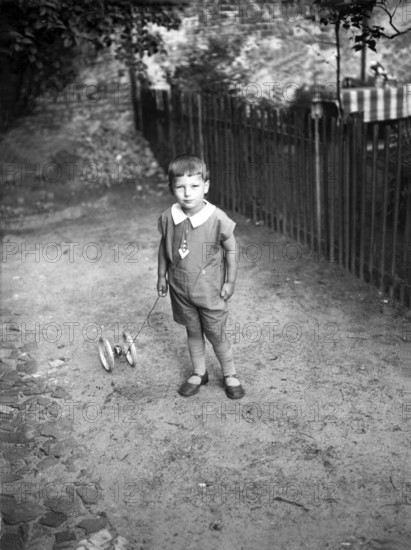 Child with toys ca. 1930s, Little boy with a toy trolley on a pavement, looking seriously into the camera, Historical photo