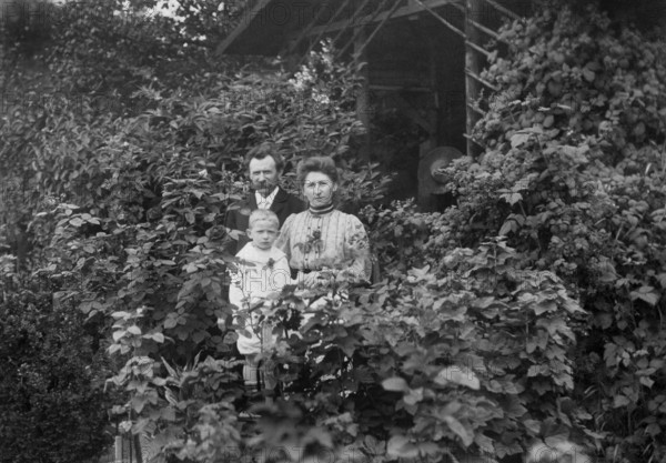 Family standing at a wooden hut, 1920s, adults with child standing in front of a house surrounded by plants, all looking serious, historical photo