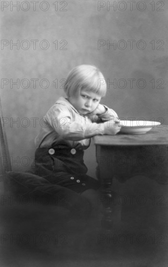 Child is angry while eating, 1920s, A boy sits thoughtfully at the table next to a bowl, Historical photo