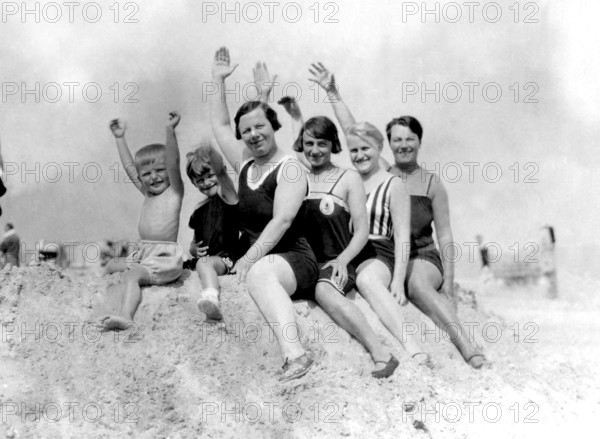 Family on a pile of sand, 1930s, group of people in swimming costumes, sitting happily on the beach, all raising their arms, historical photo