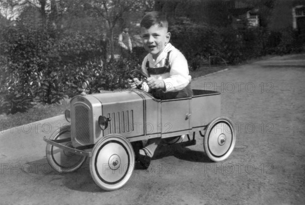 Child driving toy car, 1930s, Smiling boy driving a toy car on a street surrounded by gardens, Historical photo