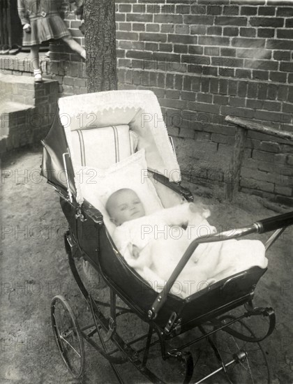 Baby in a pram, 1930s, A baby lies peacefully in a well-padded pram outside a brick building, Historical photo