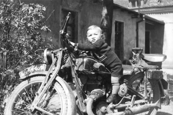 Child on a motorbike, 1930s, Small child sitting smiling on an old motorbike in a rustic setting, Historical photo