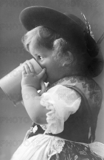 Girl drinking from a beer mug, 1930s, Small child in traditional traditional costume drinking from a mug and wearing a hat, Historical photo