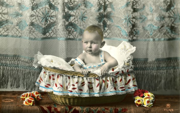 Baby sitting in basket, 1905, A baby sits in a decorative basket draped with colourful fabric, Historical photo