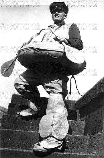 Man with boat and paddle 1940, A man in special rescue equipment stands on a staircase in front of a river, Historical photo