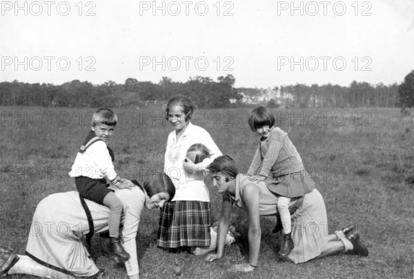 Two children riding on the backs of their mothers, 1930s, Children playing in the meadow with two adults in a cheerful atmosphere, Historical photo