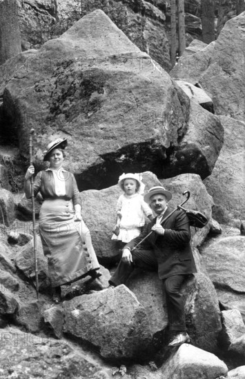 Couple with 1 child hiking, 1910, A family on a rocky outcrop in hiking clothes, holding hats and walking sticks, Historical photo