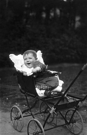 Laughing child in pram, 1910, A cheerful toddler in an antique pram in a park, Historical photo