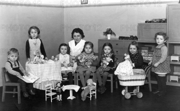 Kindergarten group toys, 1930s, children with woman in the room, playing with dolls and toys at a small table, historical photo