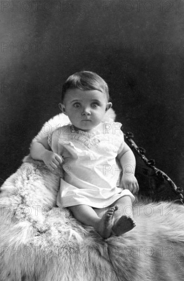 Baby sitting on polar bear fur, 1910, A baby sits on a fur, looking directly into the camera, surrounded by a black and white background, Historical photo