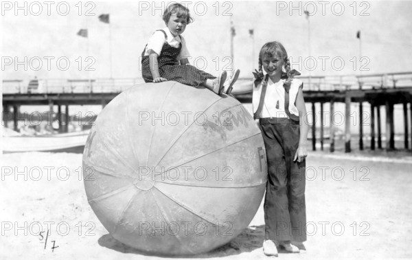 Big ball with 2 children, 1920, Two children on the beach, one sitting on a big ball, both smiling into the camera, Historical photo