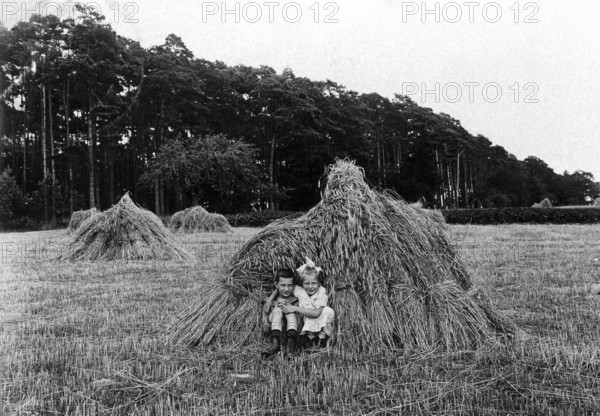 2 children in a haystack, 1920, Two children sitting in a field in front of a large bale of straw, surrounded by nature, Historical photo