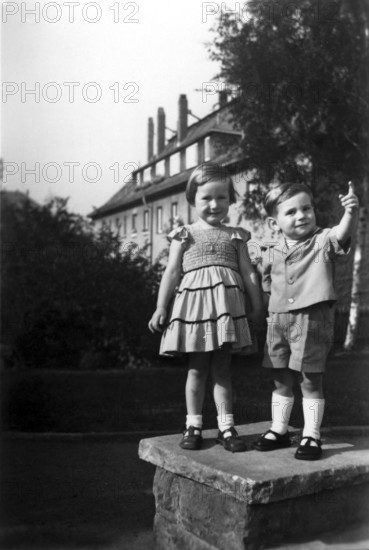 Boy showing girl something, 1920, Two children on a piece of wall outdoors with a building in the background, Historical photo
