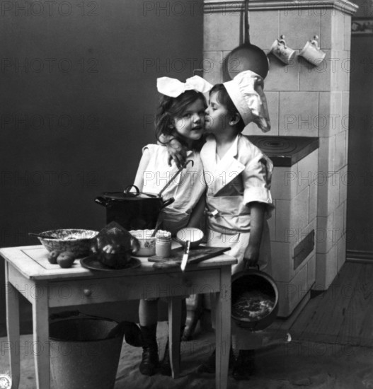 Boy kissing girl in kitchen, 1920, Two children in a kitchen setting with pots and crockery, one dressed as a cook, Historical photo