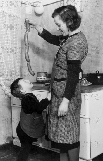 Mother feeding sausages to her son, 1930s, A woman shows sausages to a happy child in a cosy kitchen, Historical photo
