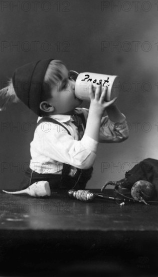Child with a beer mug, 1930s, A boy with a cap drinks from a beer mug with 'Cheers' written on it, historical photo