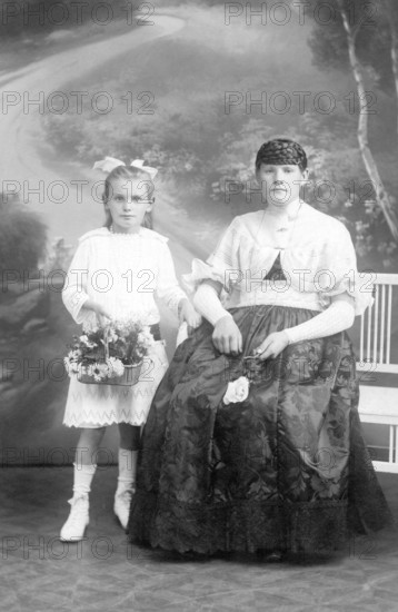 Mother and child, 1930s, A girl in a white dress holding flowers, next to a seated woman in a velvet dress, Historical photo