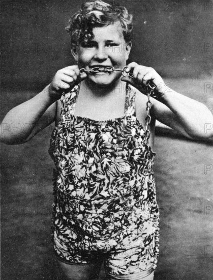 Child biting chains, 1910, A boy in a suit shows strength by pulling a chain and smiling, Historical photo