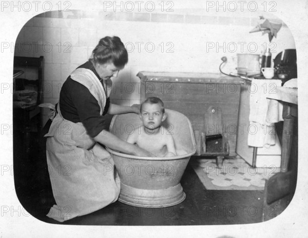 Woman bathing a boy in a small iron bathtub, 1920s, Mother bathing her child in a large tub in a nostalgic kitchen, historical photo