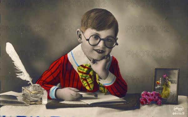 Little boy with colourful clothes sitting at desk and writing love letter, 1920s, Child with glasses in colourful clothes at desk, with quill and flowers, historical photo