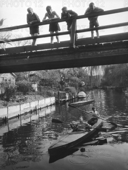 Four children on a bridge, 1930s, Children standing on a bridge while teenagers paddle underneath in a canoe, historical photo