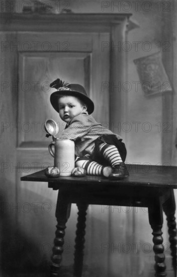 Baby traditional traditional costume beer mug, 1930s, child in traditional dress sitting on a table next to a large mug, historical photo