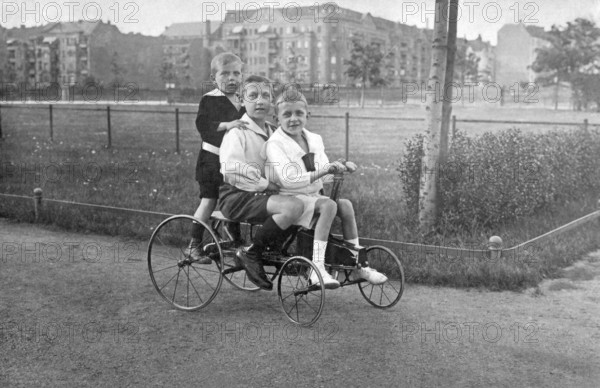Three children on a historical bicycle, 1910s, Three children riding a tricycle on a cobbled street, Historical photo