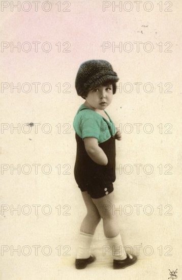 Little boy, 1920s, child with cap and green clothes in nostalgic pose in front of light background, historical photo