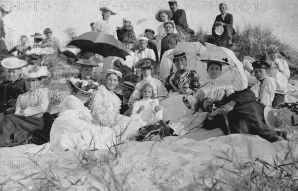 Large group of holidaymakers sitting in dunes, 1910s, Group of woman and men sitting on a sunny beach, Historical photo