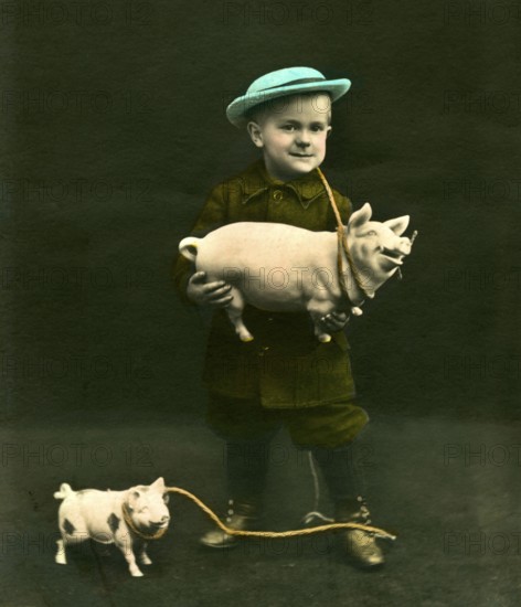 Boy with pigs, 1910s, A boy with a blue hat holds a toy sow and smiles for the photo, Historical photo