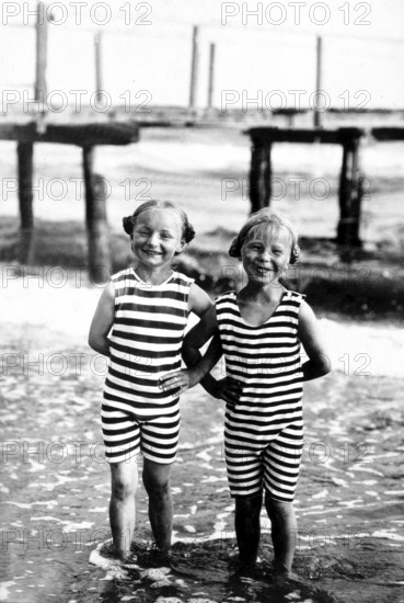 Two girls, striped swimming costume, swimwear, 1920s, Two children in striped swimming costumes on the beach, smiling in the water, wooden constructions in the background, Historical photo