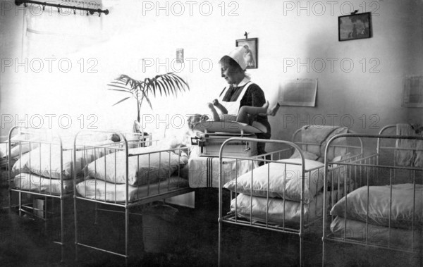 Beds Midwife cradling child, 1930s, Nurse caring for a baby in a room with several cots, Historical photo