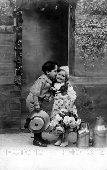 Boy kisses girl, 1920s, A boy kisses a girl on the cheek, surrounded by flowers and pewter cans, Historical photo