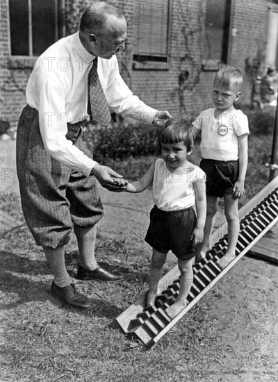 Children's physiotherapy, ca. 1925, A man helps two children on an outdoor slide in a black and white photograph, historical photo