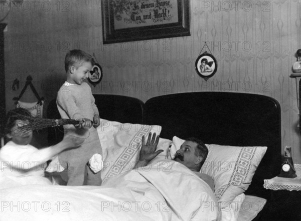 Children teasing father, 1920s, Children playing with a man in bed while one smiles and the other holds a rod, historical photo