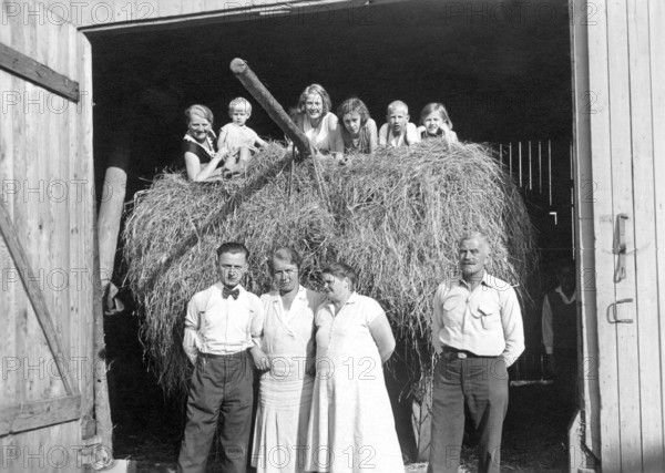 Farming family in front of the barn, ca. 1930, Family posing smiling in front of a bale of hay in an open barn, historical photo