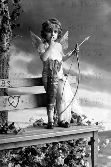 Little boy as Cupid, 1920s, A child dressed as Cupid, with wings and bow, standing on a bench, Historical photo